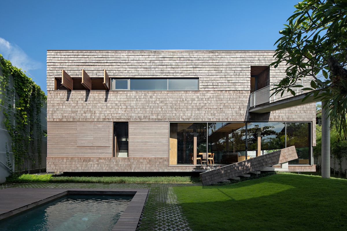 The villa's exterior features a modern minimalist design with wooden paneling. Large glass windows and a spacious patio overlook a small pool surrounded by manicured grass. A section of the building extends towards the pool, creating a unique architectural element.
