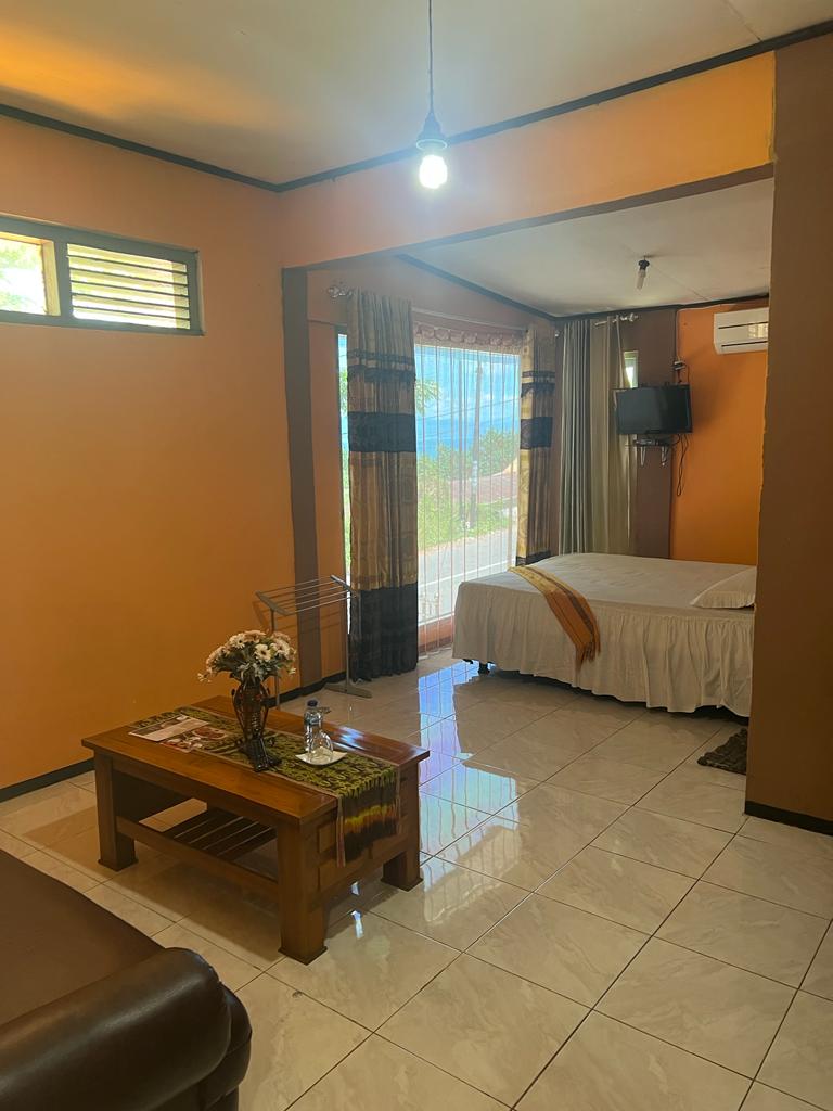 A well-lit living space features warm orange walls and glossy tiled floors. A spacious bed is positioned near a window with sheer curtains, allowing natural light to enhance the room. A small wooden coffee table holds decor and is complemented by a cozy seating area.