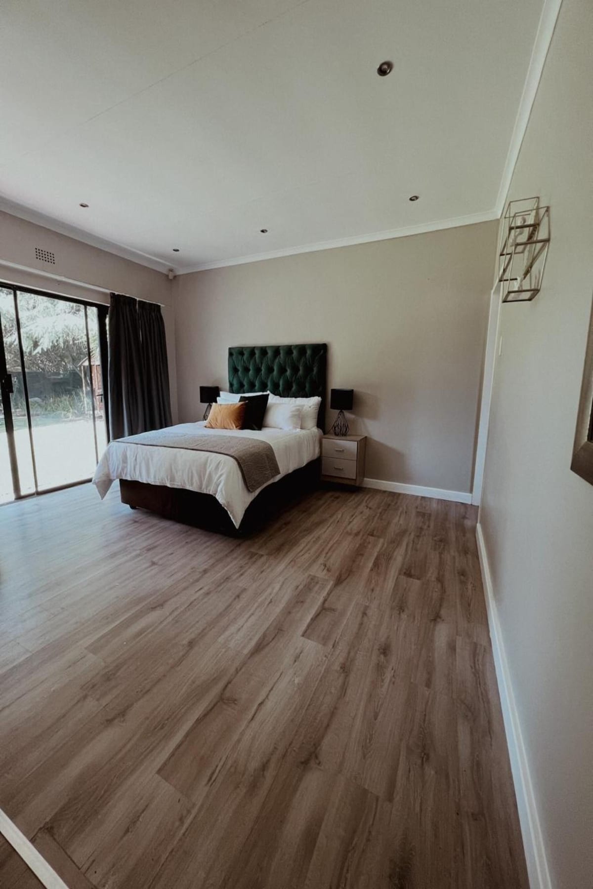 A bright bedroom features a spacious bed with a green upholstered headboard. Two bedside tables with sleek lamps are positioned on either side of the bed. Large glass doors provide access to the outdoor area, while light hardwood flooring enhances the room's open feel.