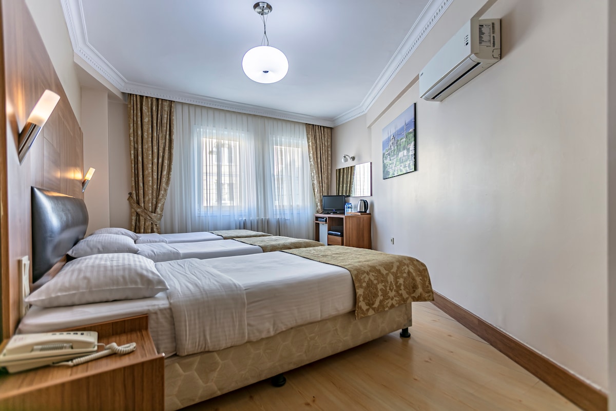 A well-lit guest room is equipped with two spacious beds dressed in crisp white linens. A wooden desk with a chair is placed near a window, which is draped with light, flowing curtains. An air conditioning unit is mounted on the wall, adding comfort to the space.