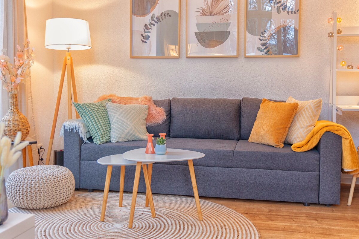 A cozy living area features a spacious gray sofa adorned with various decorative cushions. A round coffee table sits in front, accompanied by a soft pouf. Light from a floor lamp enhances the atmosphere, while framed wall art adds a touch of style.