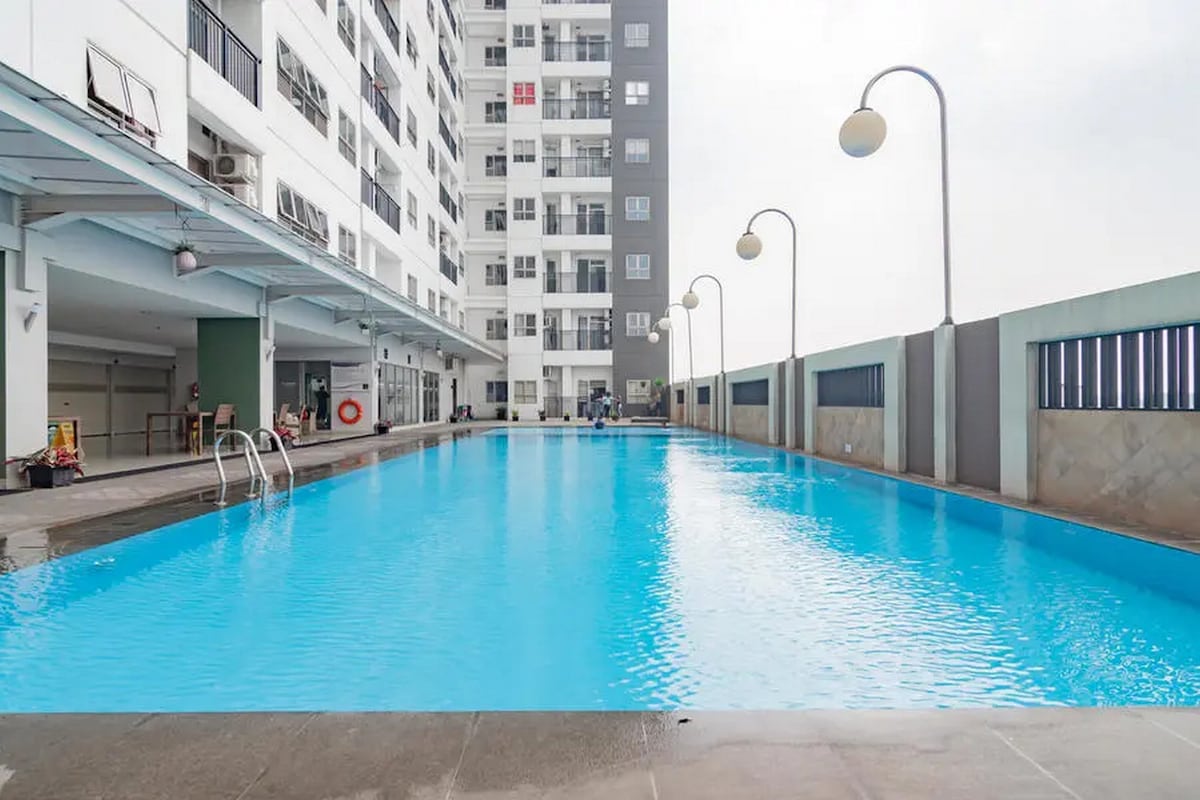 A large swimming pool extends alongside the building, featuring clear blue water. The surrounding area is well-lit with mounted lamps and includes a poolside deck. The tall building and several apartment balconies are visible in the background, contributing to a clean, modern setting.