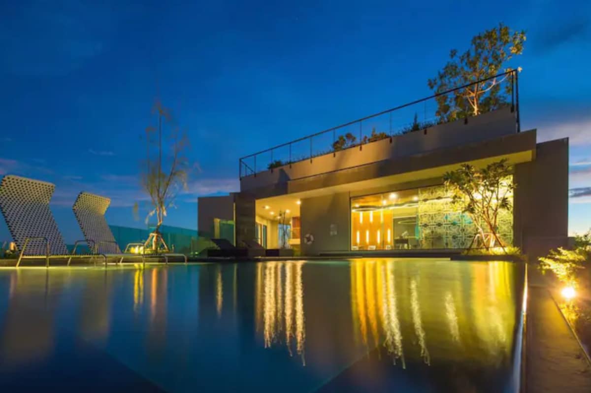 The rooftop swimming pool is illuminated under the evening sky, reflecting warm lights from the surrounding area. Modern lounge chairs are positioned beside the pool, with greenery softly framing the space, creating a serene ambiance for relaxation.