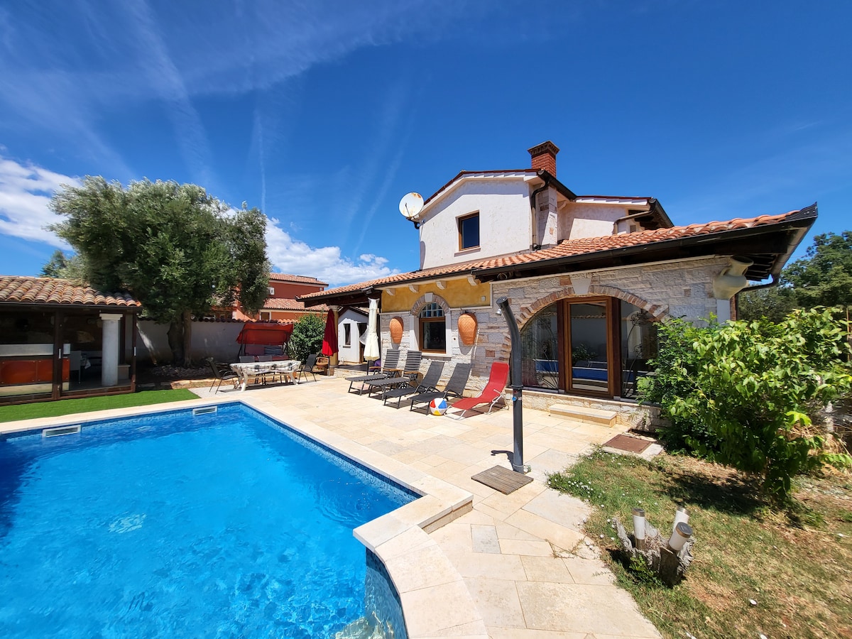 The outdoor area features a rectangular pool surrounded by sun loungers, offering space for relaxation. A stone house with arched windows and a tile roof stands in the backdrop, framed by greenery and a clear blue sky, enhancing the tranquil ambiance.