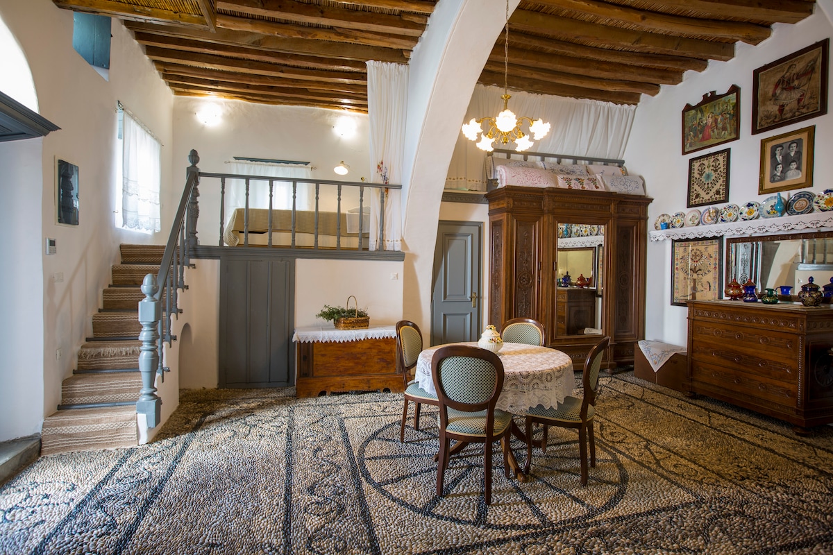 The main room features a traditional dining table surrounded by four chairs, set against a backdrop of stylish cabinetry and decorative wall art. A grand arch highlights the space, with wooden beams across the ceiling creating a warm ambiance. Soft natural light filters through the windows.
