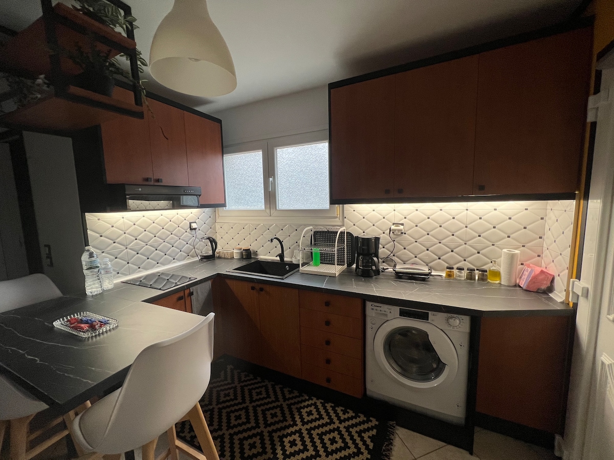 A compact kitchen is equipped with dark wooden cabinets and a modern countertop. A washing machine is located next to the sink, and small appliances are arranged on the counter. Natural light enters through a window with textured glass, enhancing the functional design.