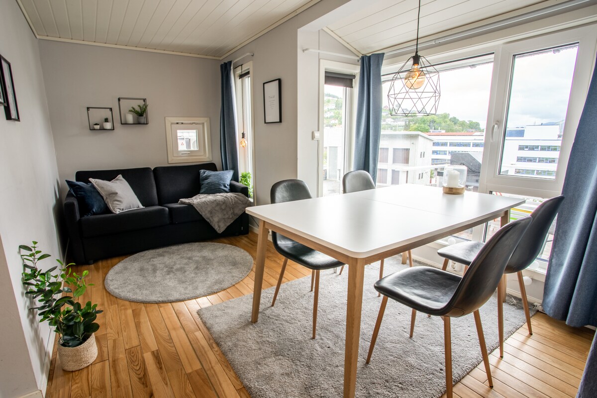 An open living area with a black sofa is positioned against a light-grey wall. A round rug lies beneath a dining table surrounded by four chairs. Natural light enters through large windows, partially covered by blue curtains, creating a bright and inviting atmosphere.