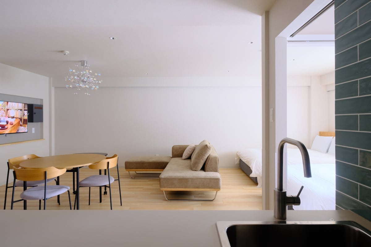 An open living area is visible, featuring a modern, light-colored sofa adjacent to a round dining table with four stylish chairs. A compact kitchen is partially seen, showcasing a sleek sink and countertop. A well-lit bedroom area is found in the background, creating an inviting space.
