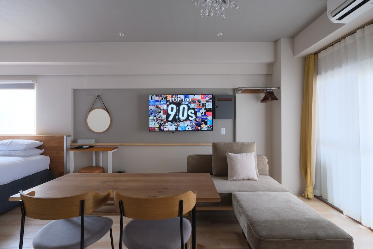 A bright lounge area features a large 55-inch flatscreen TV mounted on the wall, displaying colorful visuals. A comfortable sofa is positioned nearby, with a simple round mirror and a wooden dining table with two chairs adjacent, while ample natural light flows from large windows.