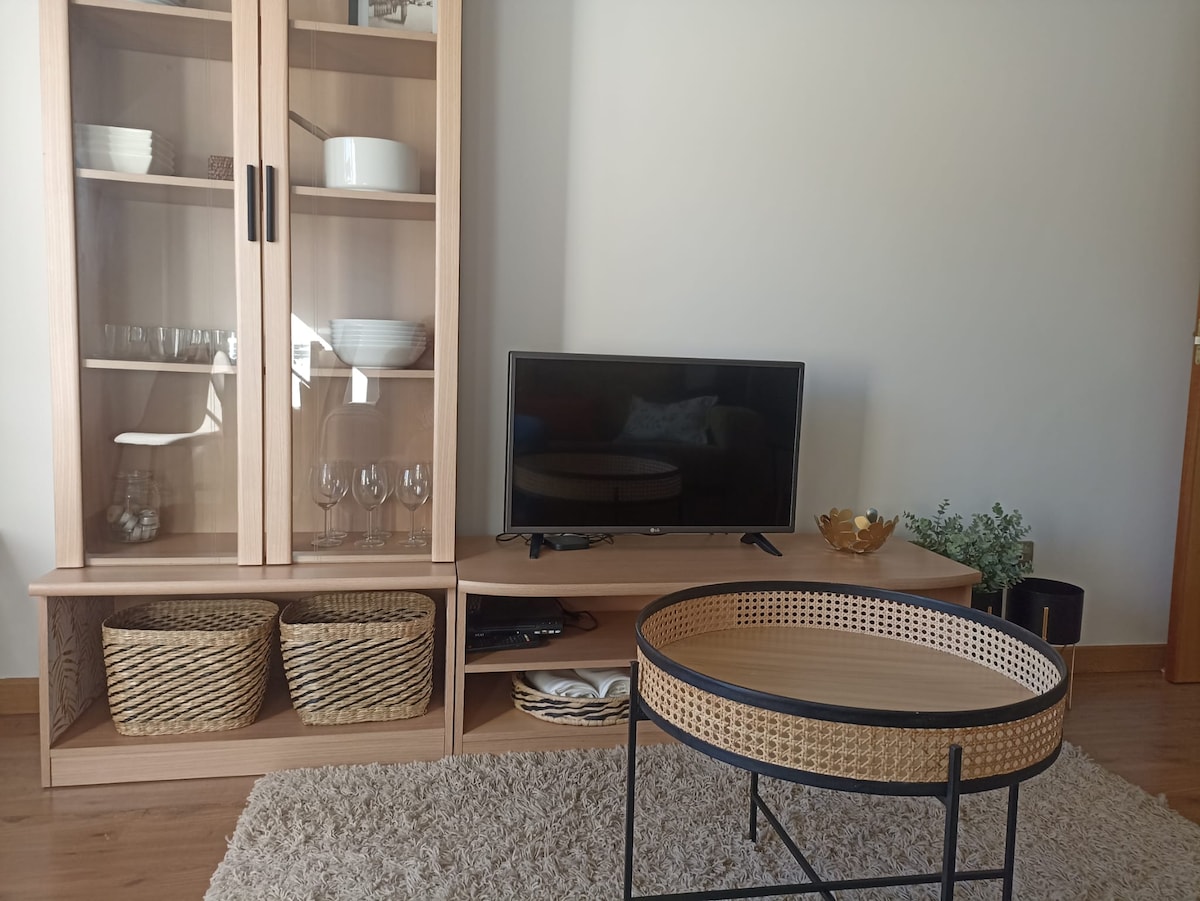 A living area is presented with a light wood cabinet displaying tableware and glassware. A television is positioned on the cabinet, accompanied by a circular coffee table featuring a woven design. Baskets are placed below for storage, and a small plant adds a touch of greenery.