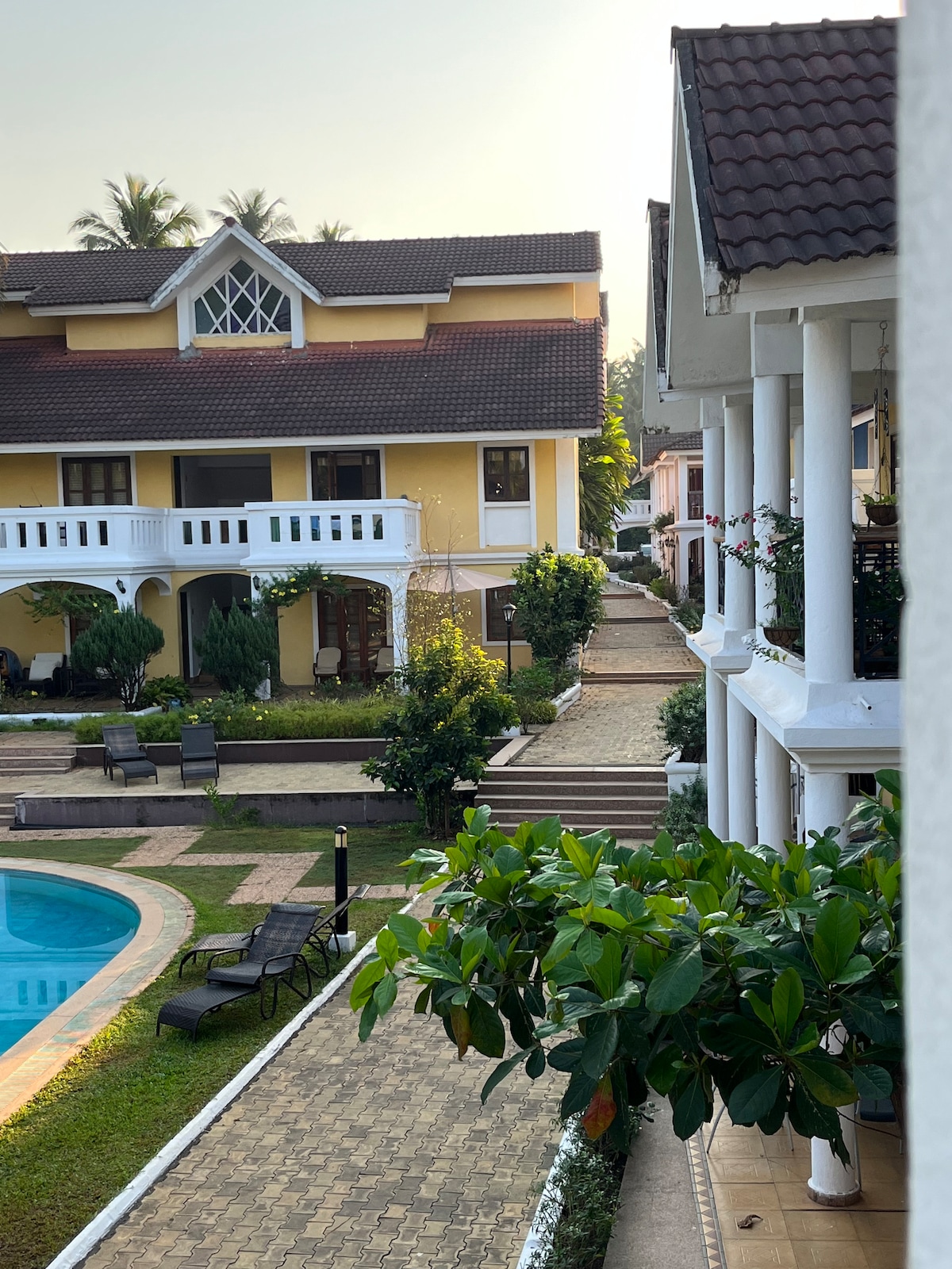 A scenic view of a tranquil resort is displayed, featuring charming buildings nestled amidst lush greenery. A swimming pool is visible in the foreground, surrounded by lounge chairs and pathways leading to the well-maintained gardens.