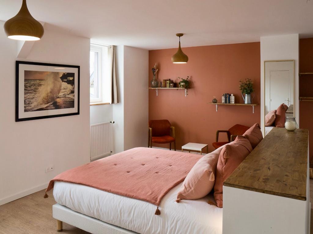 A spacious bedroom features a queen size bed adorned with soft pillows and a textured blanket. A warm accent wall adds colour, while a photograph of ocean waves emphasizes the decor. A small seating area is present, alongside a minimalist shelf showcasing decorative items.