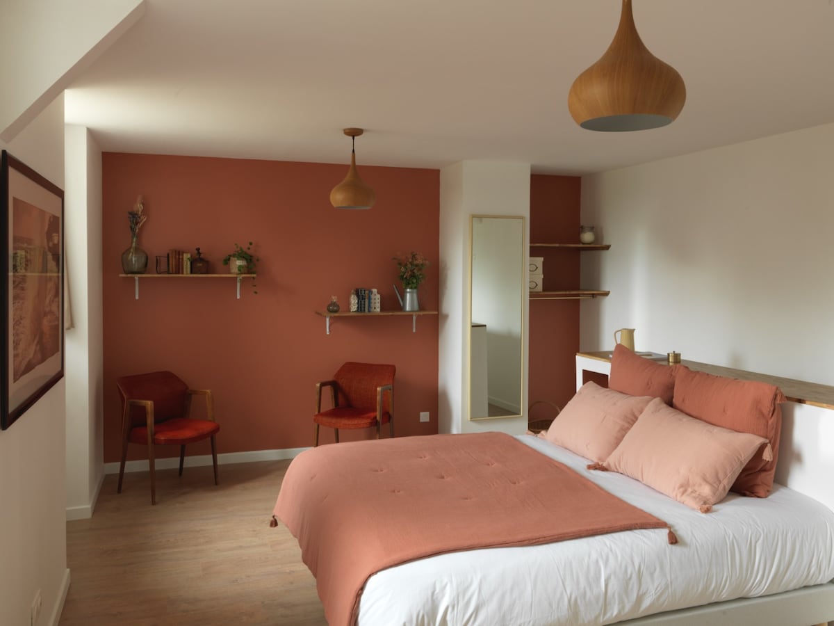 A spacious bedroom features a queen-size bed adorned with soft, warm-toned bedding. Two comfortable chairs are positioned nearby, and a small shelf showcases decorative items and plants. The room is illuminated by a pendant light, complementing the warm-colored accent wall.