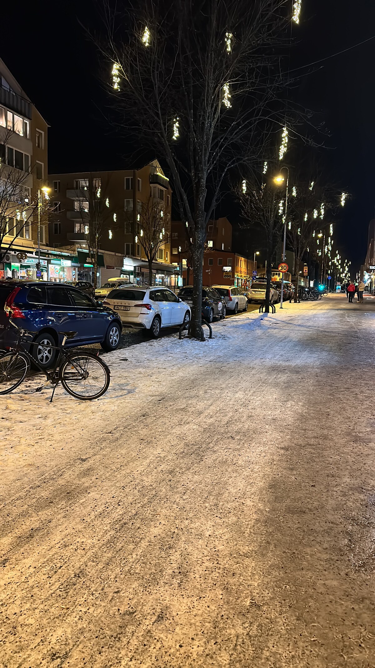 A snowy street scene is captured at night, featuring parked cars lined along a well-lit avenue. Tree branches are adorned with sparkling lights, illuminating the pathway. A bicycle rests nearby, complementing the neighborhood's serene winter atmosphere.