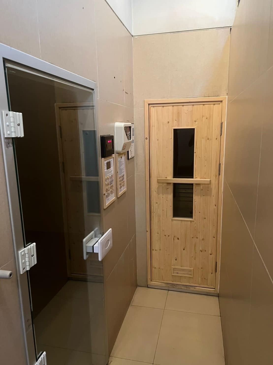 A wellness area is shown with a wooden sauna door featuring a vertical window. Adjacent is a glass door leading to a steam room, both set against tiled walls and a neutral floor. Information and controls are displayed on the wall beside the sauna.