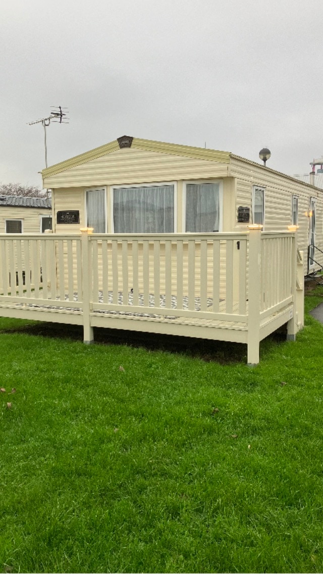 A charming caravan is situated on well-maintained green grass, featuring a wraparound wooden deck. The exterior showcases light-colored siding and large windows, offering a welcoming appearance. Soft lighting enhances the deck area, providing a cozy spot for relaxation.
