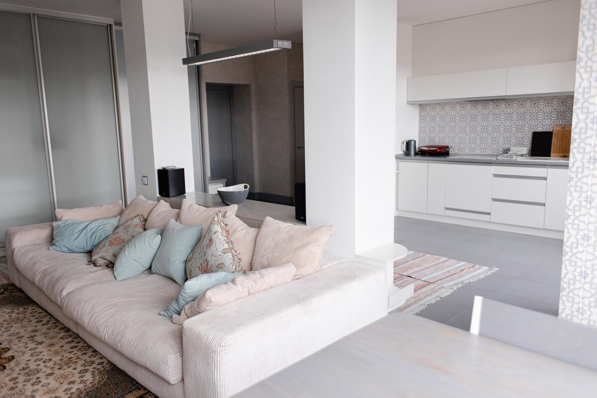 A spacious living area features a light-colored sofa adorned with several soft pillows. The open kitchen is visible in the background, showcasing modern cabinetry and a patterned backsplash. Natural light fills the space, enhancing the clean and contemporary design.