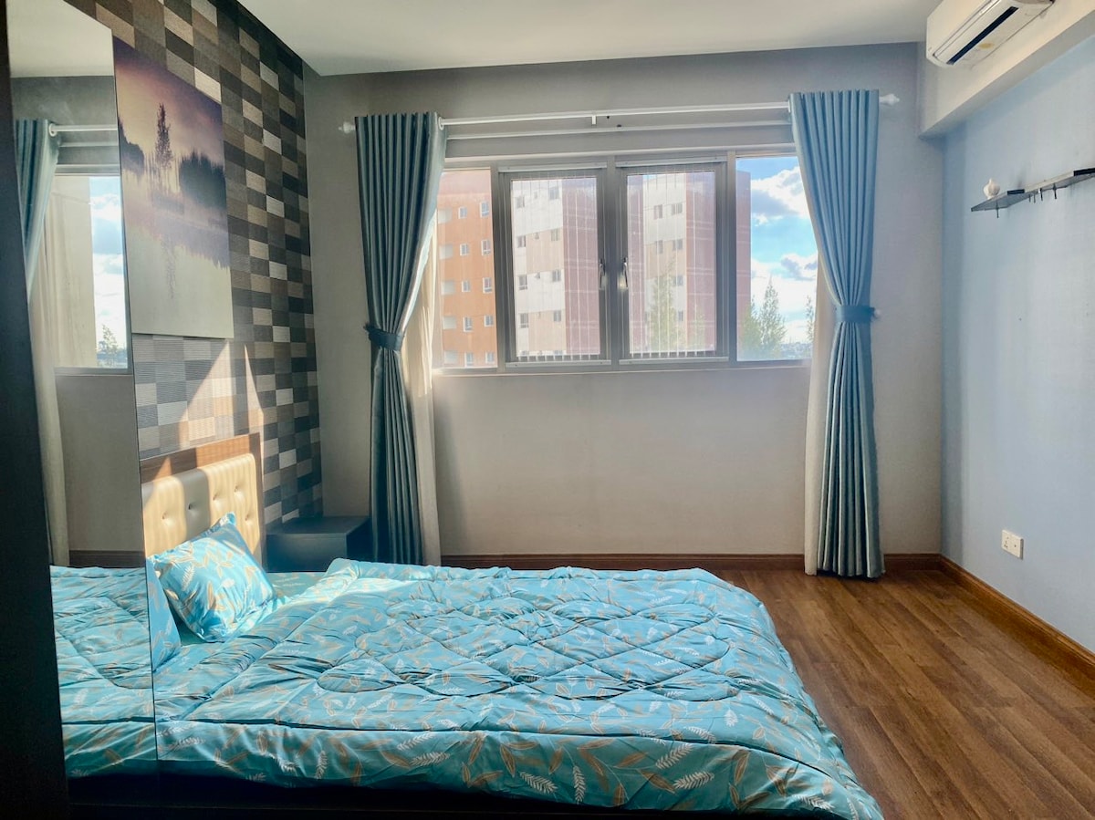 A serene bedroom is presented with a large bed adorned with a blue patterned quilt. Natural light floods through two windows, complemented by light curtains. The walls feature a mix of neutral tones and textured patterns, enhancing the overall spacious feel of the room.