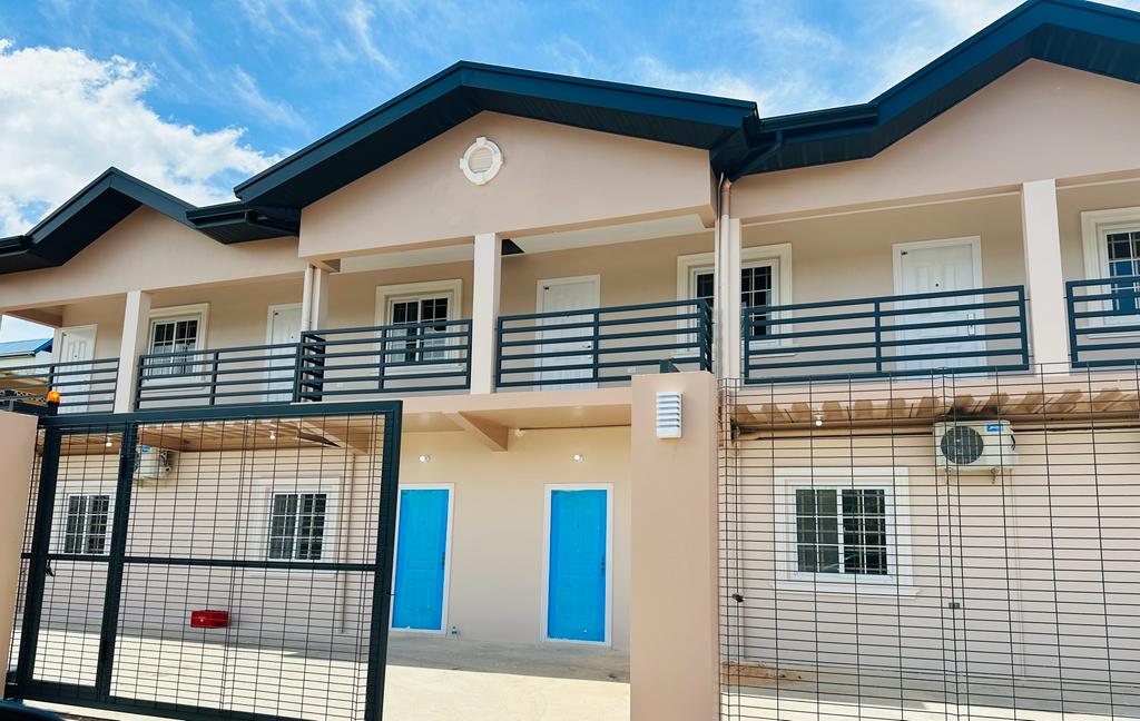 The exterior of a contemporary building features a gated entrance with a secure metallic fence. The structure is painted in neutral tones, adorned with two distinct blue doors, and highlighted by large windows on the upper level, creating an inviting and fresh appearance under a bright sky.
