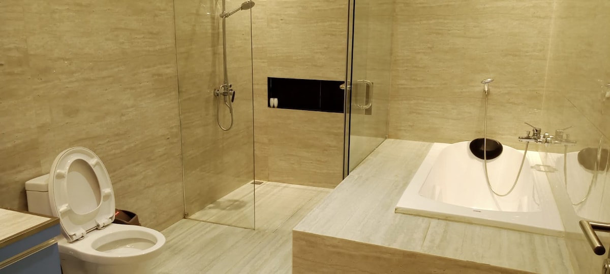A spacious bathroom features a modern glass shower and a sleek bathtub integrated into the design. Neutral-toned stone walls and flooring create a calming atmosphere. A toilet is positioned to the left, enhancing the room's functionality.