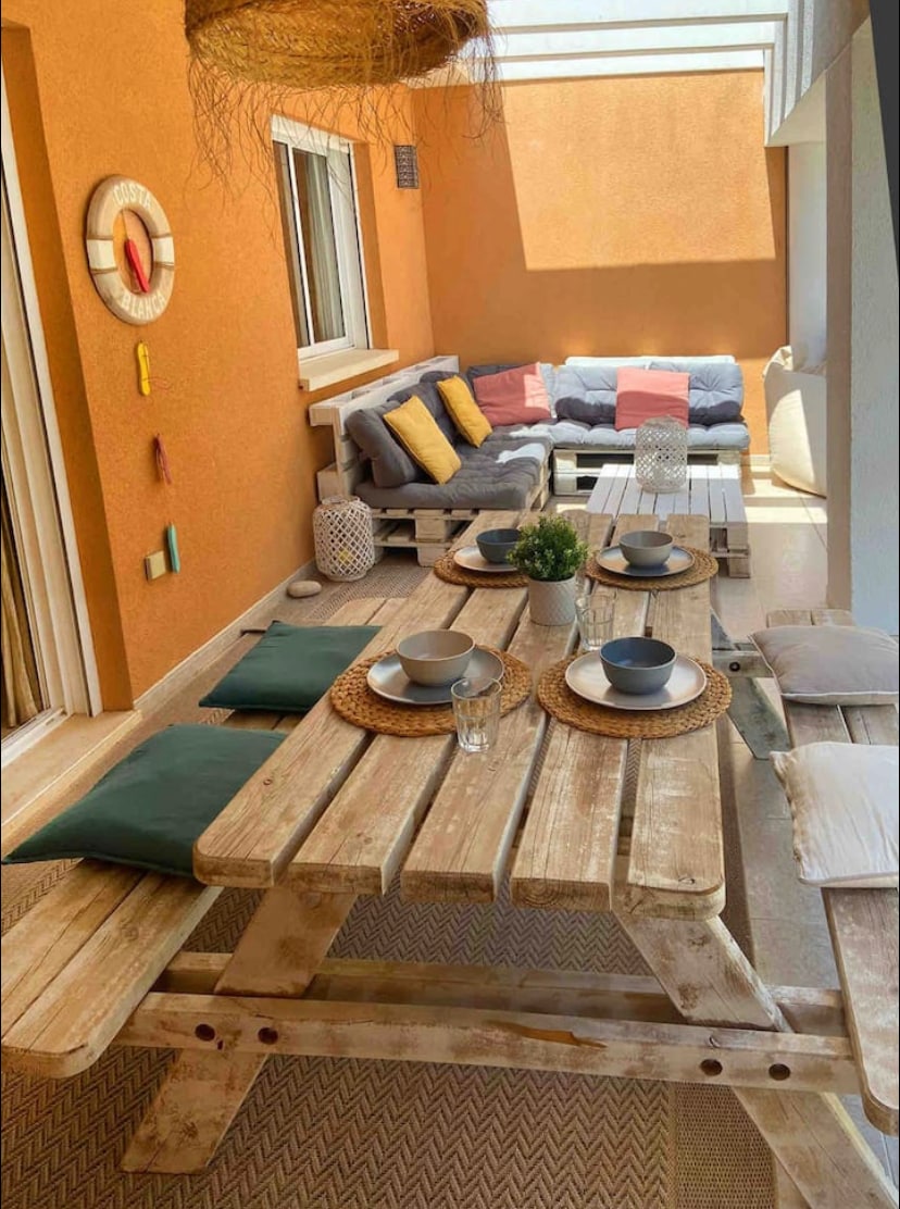 A shaded terrace features a rustic picnic table set with plates and glasses, complemented by decorative placemats. A cozy seating area includes a cushioned sofa arranged with colorful pillows. The wall is painted in warm orange, adding a vibrant touch to the space.