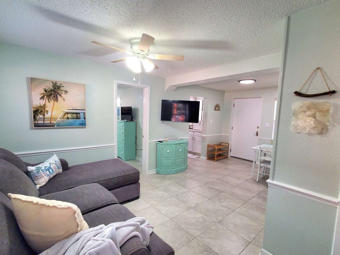 A comfortable living area features a sectional sofa adorned with a throw, complemented by light green walls. A ceiling fan adds functionality and a framed artwork depicting a classic car and palm tree enhances the space. A wall-mounted TV is visible, providing entertainment options.