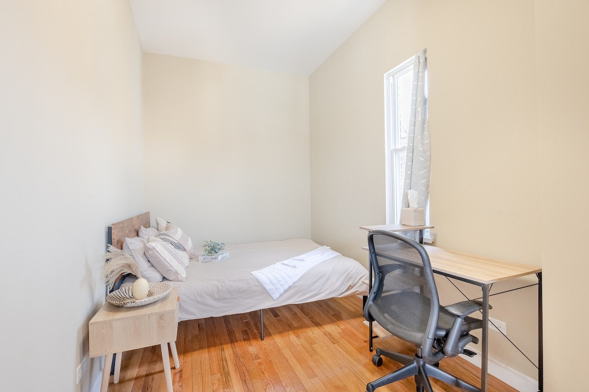 A bright bedroom features a cozy queen bed covered with neutral linens and accented by decorative pillows. Natural light enters through a window, illuminating the warm wooden floor. A desk and chair offer a designated workspace alongside a simple nightstand.
