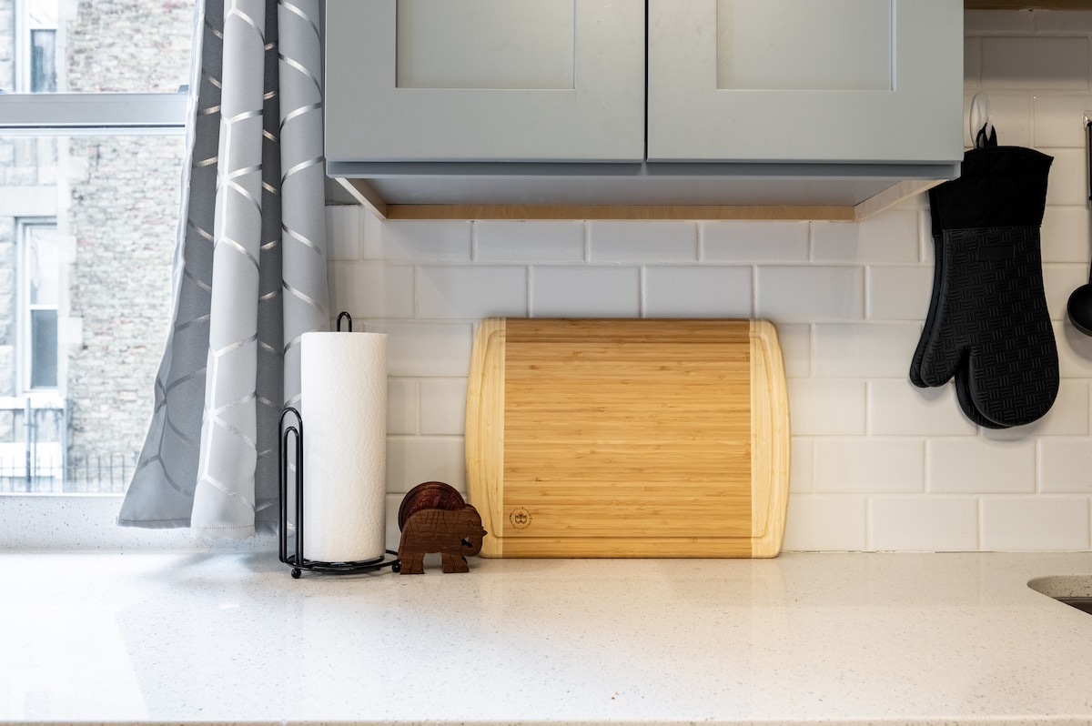 A clean and modern kitchen counter is presented with a bamboo cutting board, a decorative wooden elephant, and a paper towel holder. Light grey curtains frame a window, while a black oven mitt hangs nearby, contributing to the organized space.