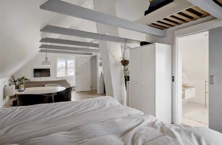 A spacious bedroom is seen with a comfortable bed covered in a white duvet. Exposed wooden beams draw the eye towards a cozy seating area with a light-colored sofa. A white cupboard stands near a doorway leading to a minimalist bathroom.