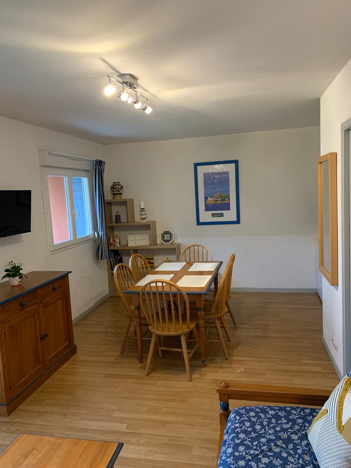 A dining area features a wooden table surrounded by six chairs, illuminated by track lighting above. A sideboard provides additional storage, while a framed artwork adorns the wall. A window offers natural light, with blue curtains partially drawn.