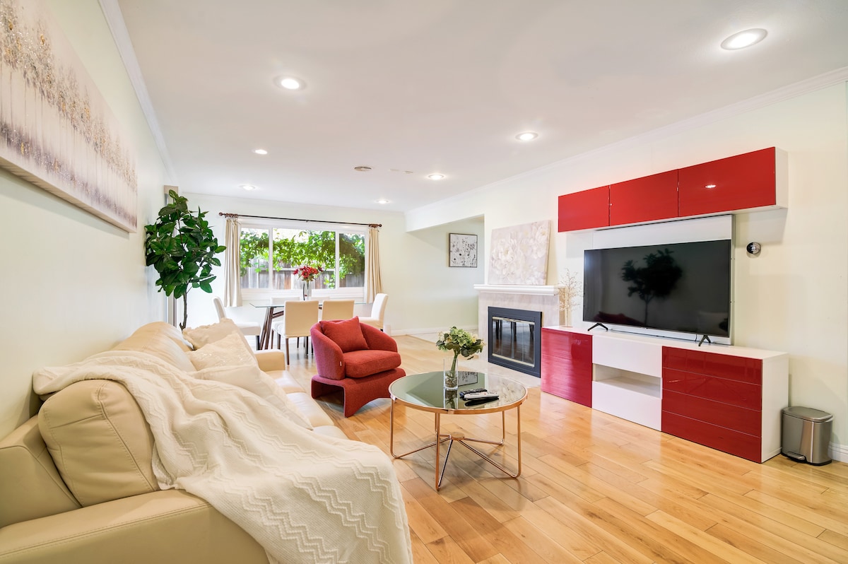 A bright living room featuring a light-colored sofa and a glass coffee table. A large flat-screen TV is mounted on a red panel cabinet, complemented by a cozy fireplace. Natural light enters through the window, illuminating the dining area with a white table and chairs.
