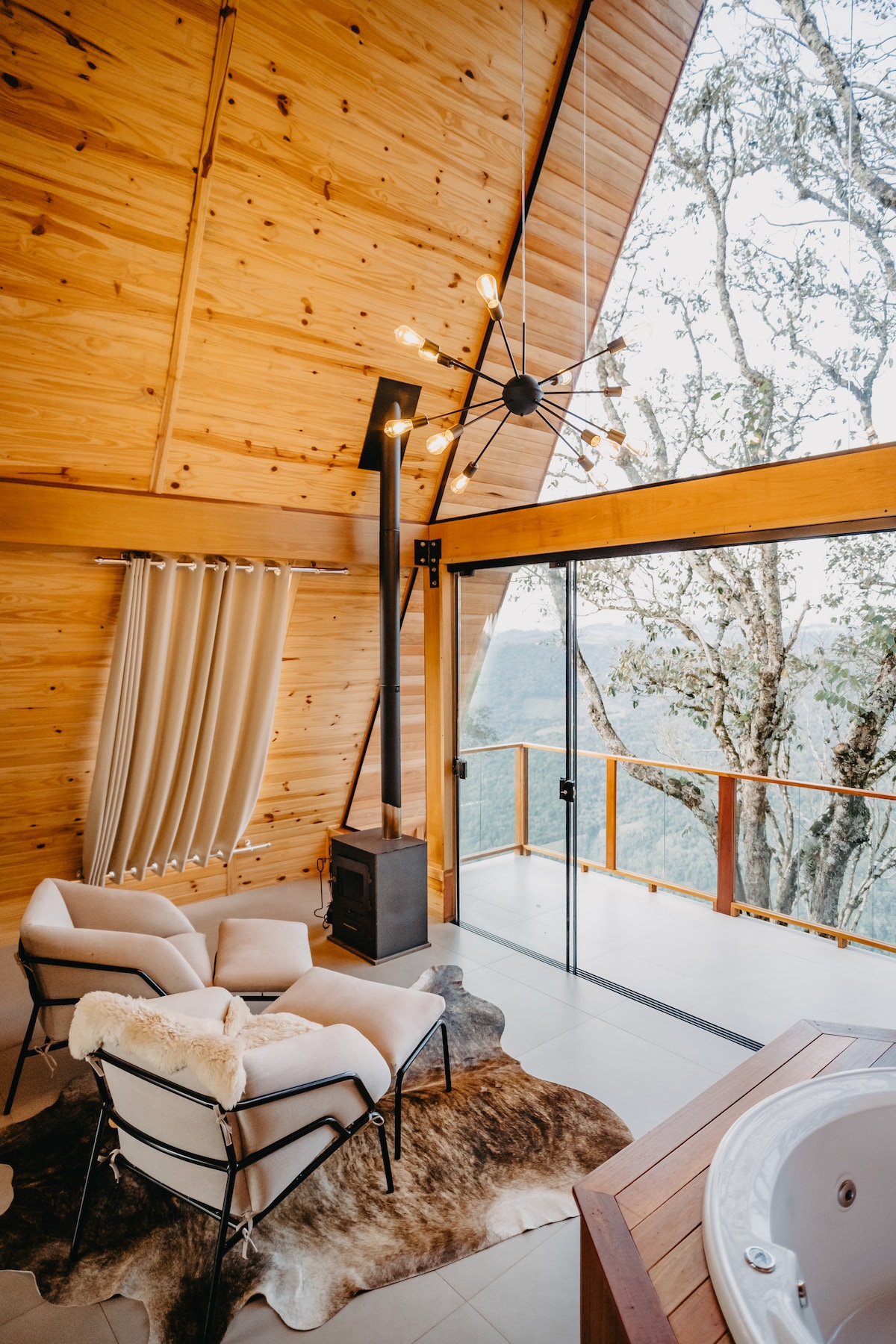 A cozy corner is featured with two armchairs positioned near a minimalist fireplace. Large glass doors provide a view of the surrounding mountain landscape, while natural wooden walls and beams add warmth to the space. A soft area rug enhances the inviting atmosphere.