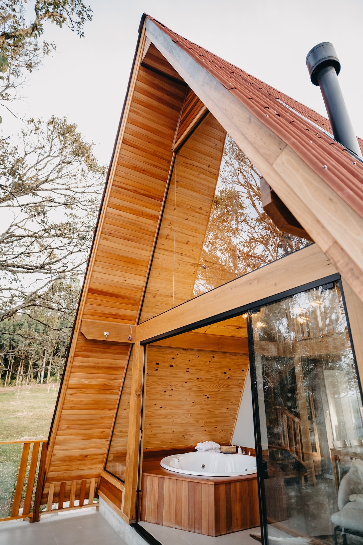 The exterior of the A-frame chalet features a striking wooden design with large glass panels. An outdoor hot tub is positioned beside the entrance, surrounded by natural greenery, enhancing the connection with the tranquil landscape.