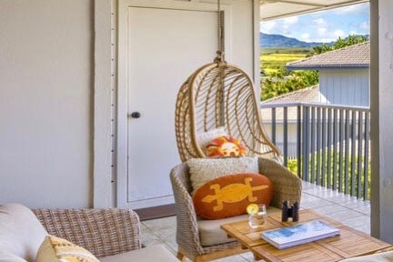 A cozy balcony area features a hanging wicker chair adorned with a decorative pillow. A small wooden table holds a refreshing drink, while serene views of green hills and neighboring buildings are visible beyond the railing.