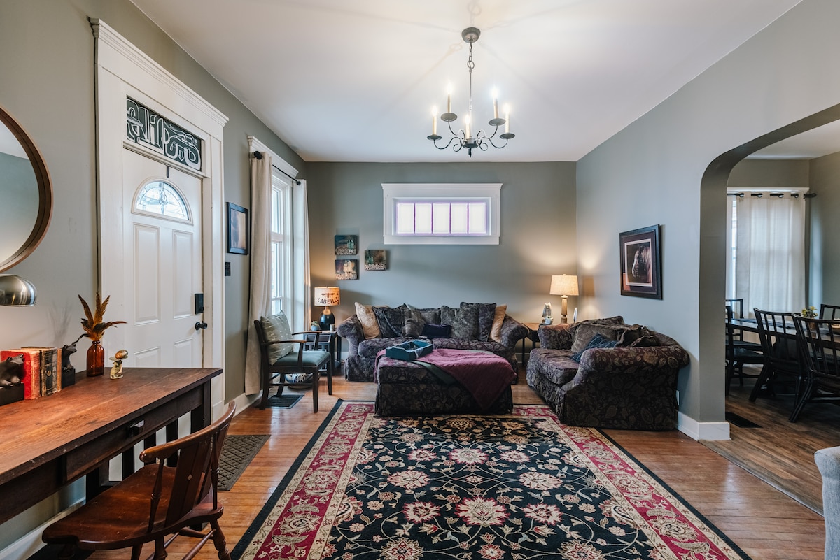 A living area is presented with a blend of modern and traditional furnishings. A large rug covers the wooden floor, while a comfortable sectional sofa and armchair provide ample seating. Natural light enters through large windows, and a chandelier adds to the room's ambiance.