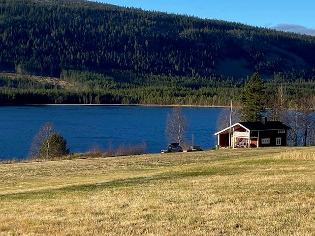 A charming house is set against a backdrop of lush green hills and a serene lake. The exterior features a mix of large windows and a spacious deck. Open grassy areas surround the property, providing a sense of tranquility and connection with nature.