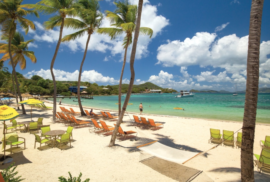 A sandy beach is bordered by swaying palm trees, with rows of colorful lounge chairs set up for relaxation. Crystal-clear waters and gentle waves are visible in the background, while boats can be spotted in the distance under a bright, blue sky.