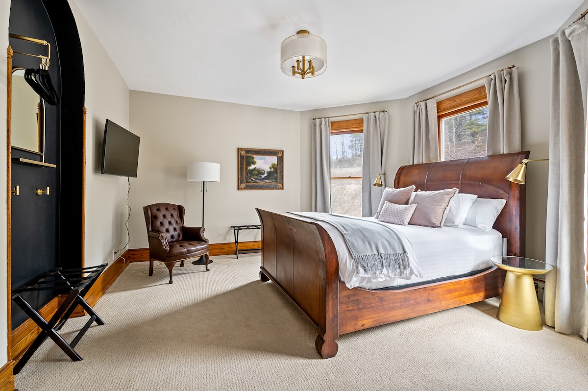 A spacious bedroom features a queen sleigh bed with crisp white bedding and decorative pillows. Natural light enters through two windows, illuminating a cozy seating area with a brown leather chair. A flat-screen TV is mounted on the wall, complemented by a stylish gold light fixture.