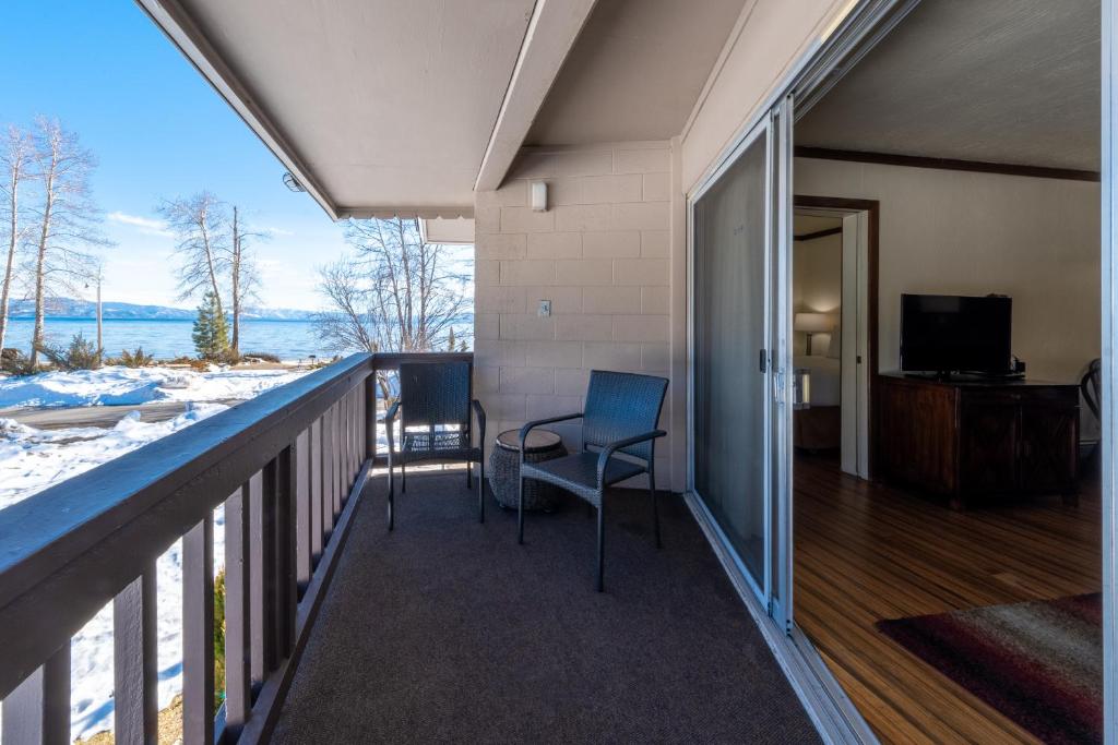 A private balcony features two metal chairs positioned on a carpeted surface. A view of the snow-covered landscape and lake is visible in the background, with tall trees lining the shoreline under a clear blue sky.