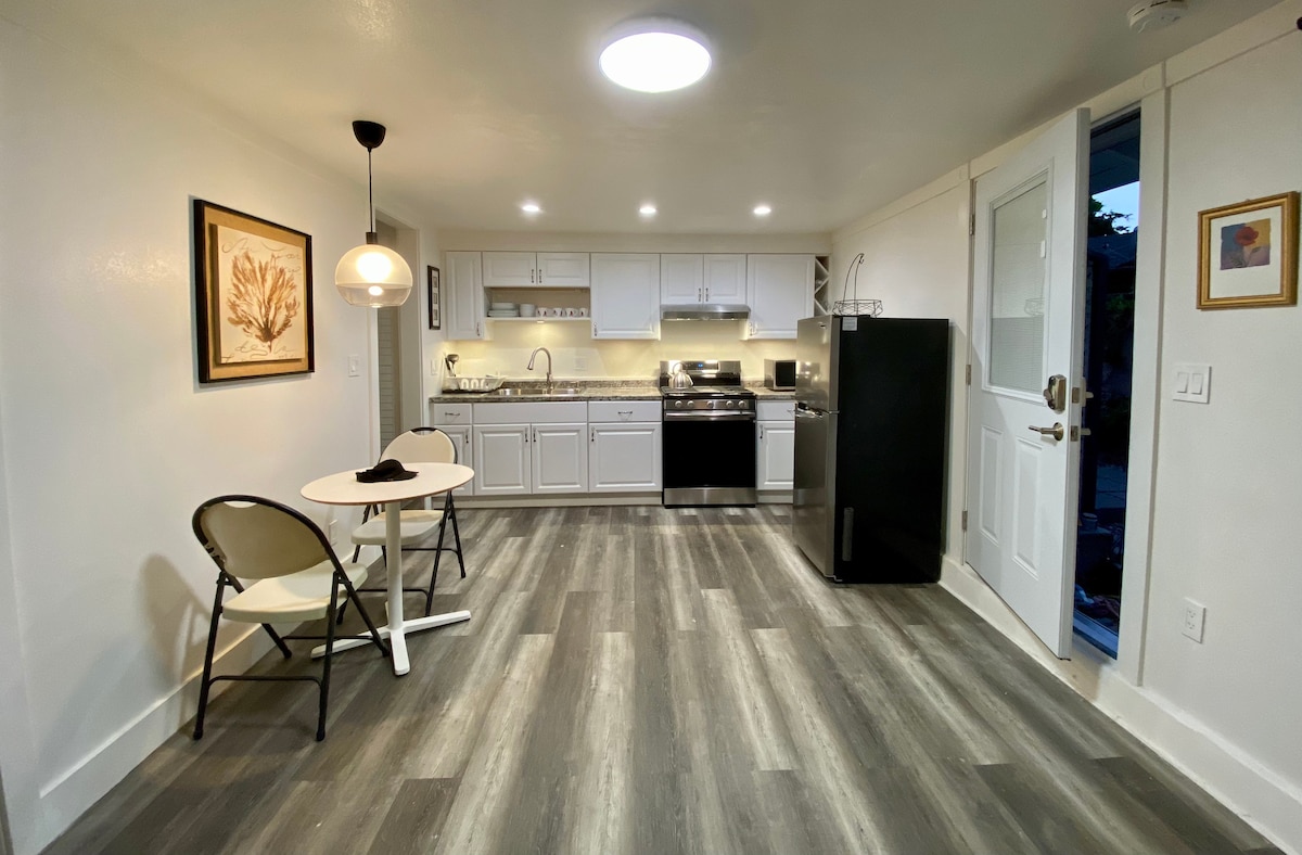 A well-lit kitchenette features modern appliances, including a stove, microwave, and refrigerator. A small dining table with two chairs provides a cozy space for meals. Light-colored cabinets and a framed artwork add a touch of charm to the clean, open layout.