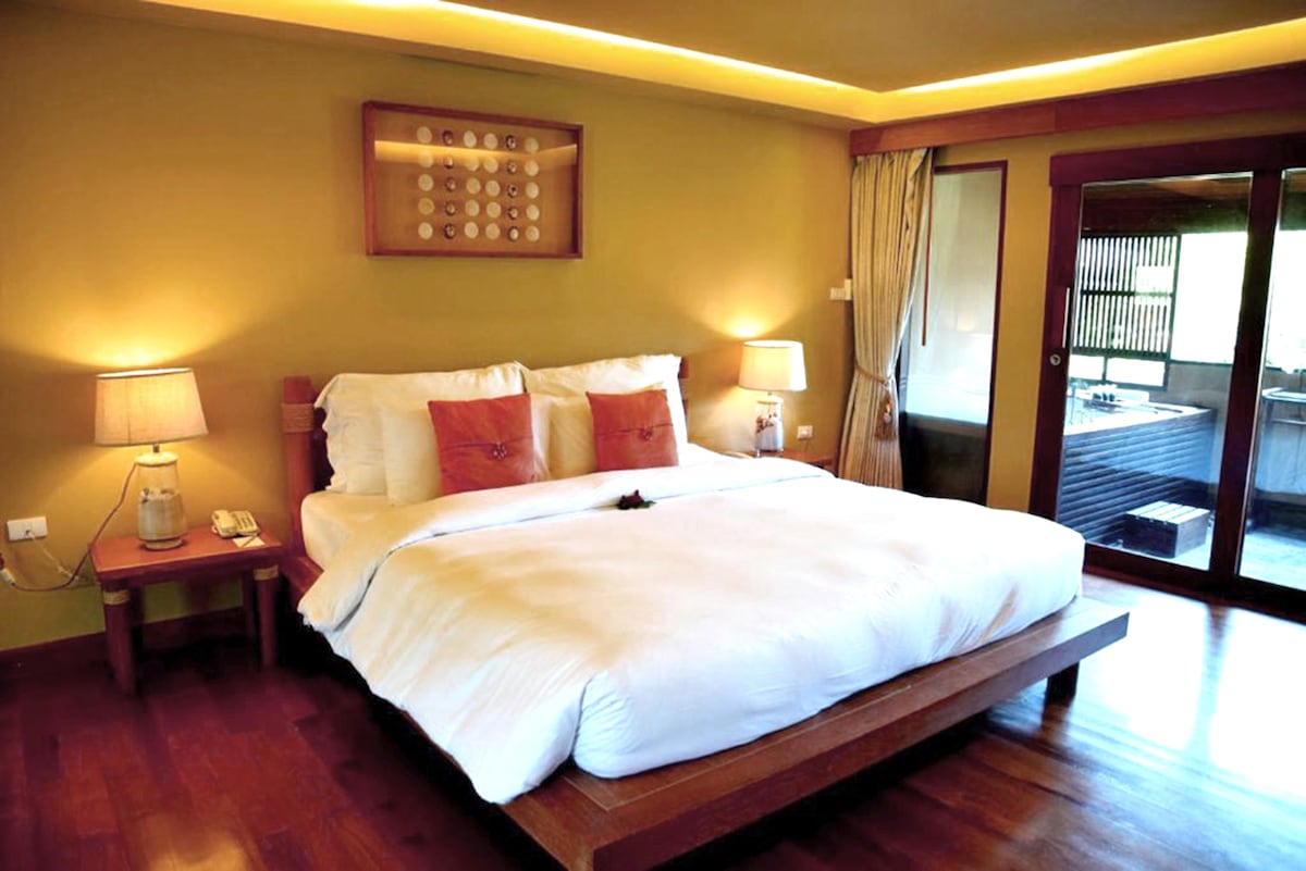 A spacious room features a king size bed with soft linens, accompanied by two bedside lamps. Natural light fills the area through large windows, revealing an ensuite bathroom with a bathtub. Warm yellow walls and wooden flooring contribute to a calming ambiance.