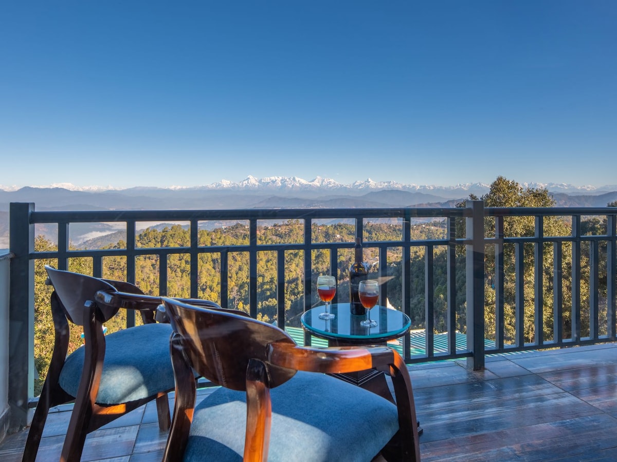 A balcony offers a stunning view of distant snow-capped mountains, framed by coniferous trees. Two wooden chairs with blue cushions face a small glass table, which holds two drinks. The clear blue sky provides a serene backdrop to this inviting outdoor setting.