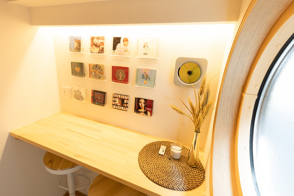 A small nook is illuminated by warm light, featuring a smooth wooden table paired with a white stool. A collection of framed artwork and compact discs is displayed on the wall above. A decorative golden plate and dried wheat are placed on the table.