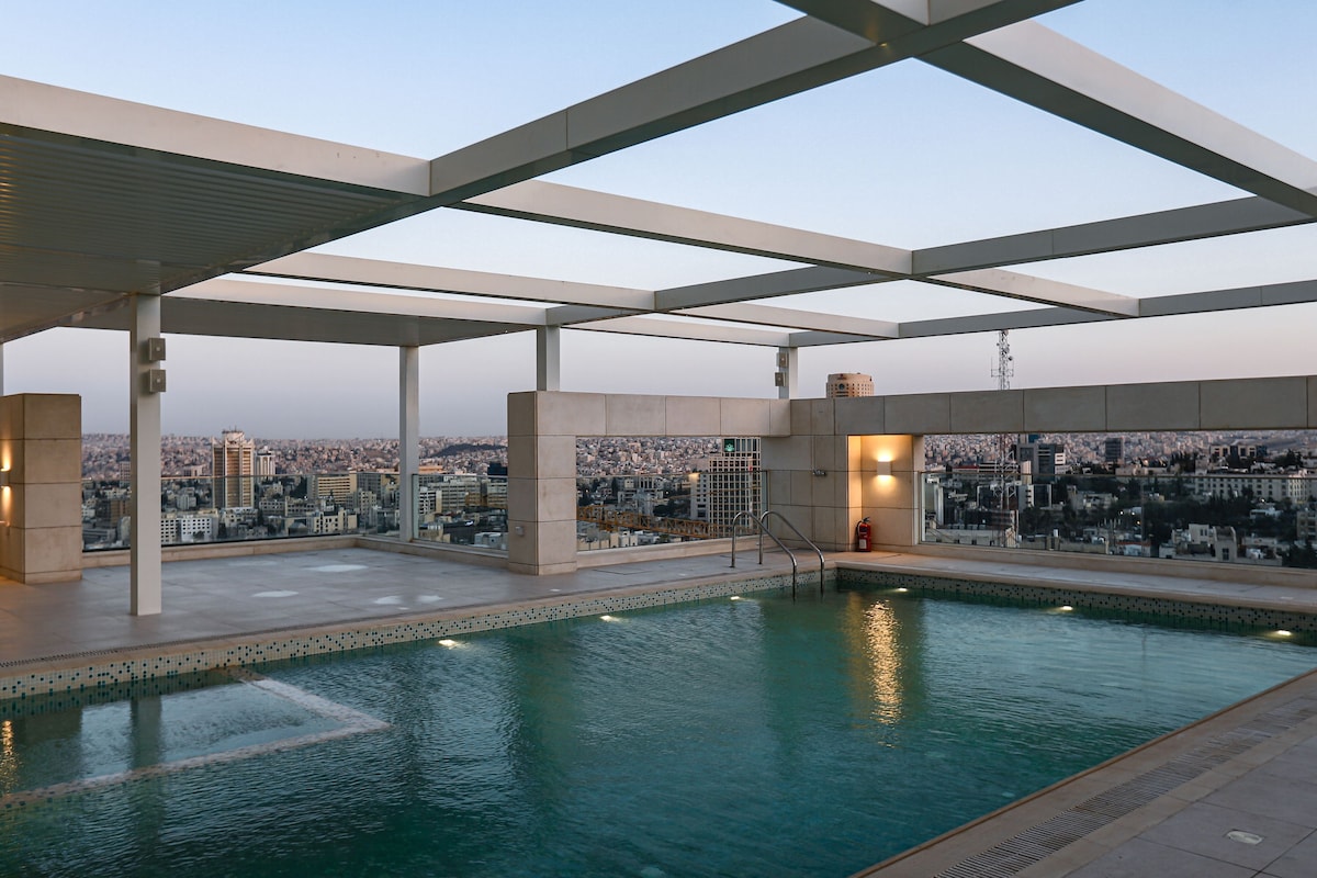 An outdoor swimming pool area features clear water framed by a modern pergola, allowing for stunning views of the city skyline. The pool is surrounded by tiled surfaces and has a ladder for easy access. The setting offers a sense of openness and tranquility.