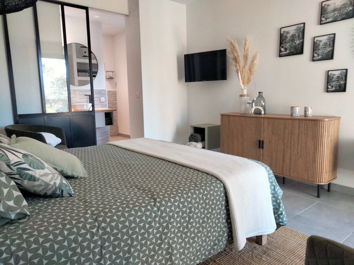 A comfortable suite features a spacious bed adorned with a patterned blanket. A minimalist side table and a sleek cabinet are visible, along with a television mounted on the wall. Natural light filters in, enhancing the contemporary decor with greenery accents.