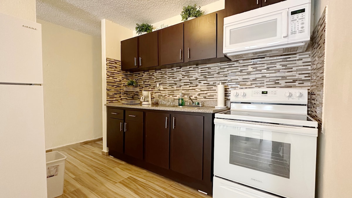 A modern kitchen is presented with dark wooden cabinetry and stylish backsplash tiles. Essential appliances are included, such as a white refrigerator, a stove with an oven, and a microwave. Natural light enhances the space, highlighting the wood-style flooring.