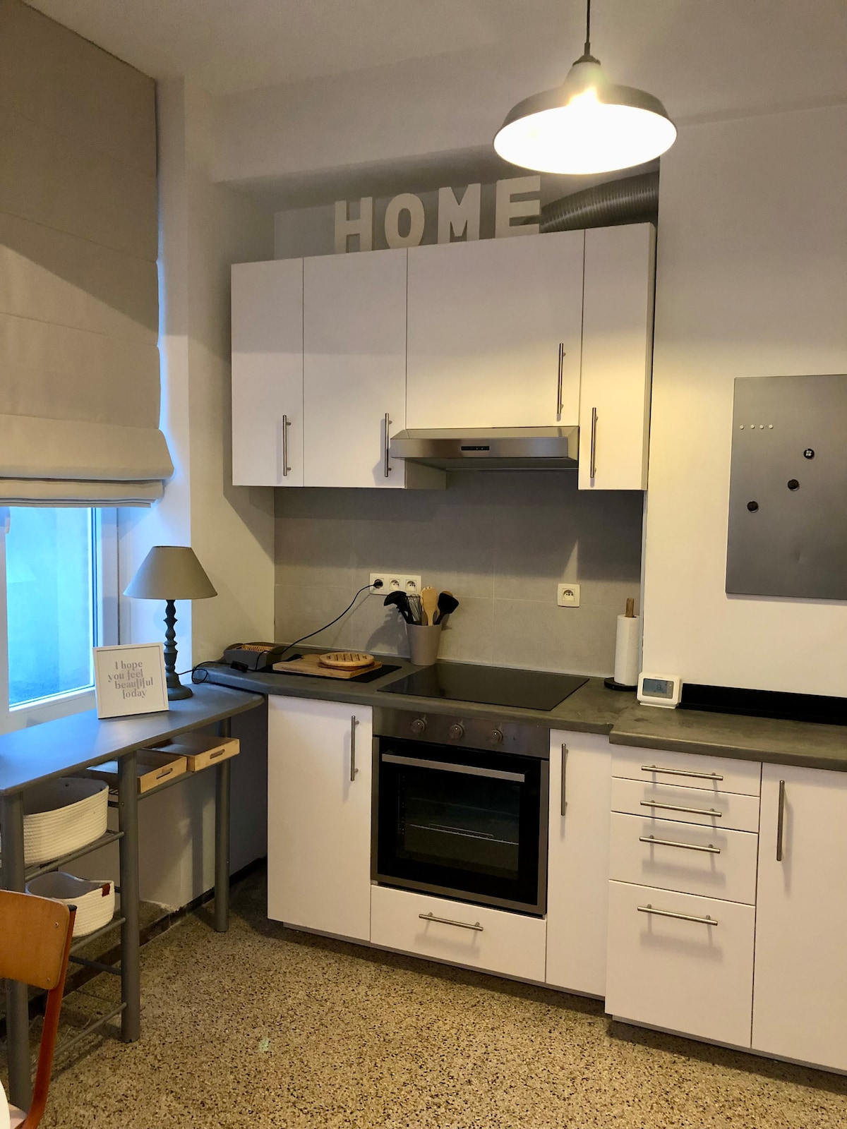 A compact kitchen is shown, featuring white cabinetry and a metallic vent hood above a built-in oven. A countertop offers space for meal preparation, accompanied by a decorative lamp and kitchen accessories. Natural light enters through a nearby window with a soft fabric shade.
