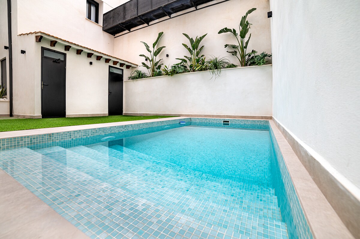 An outdoor swimming pool features clear, inviting water surrounded by modern tile work. Lush green plants provide a backdrop against the white wall, creating a serene atmosphere. Two dark doors, likely leading to private spaces, complement the setting.