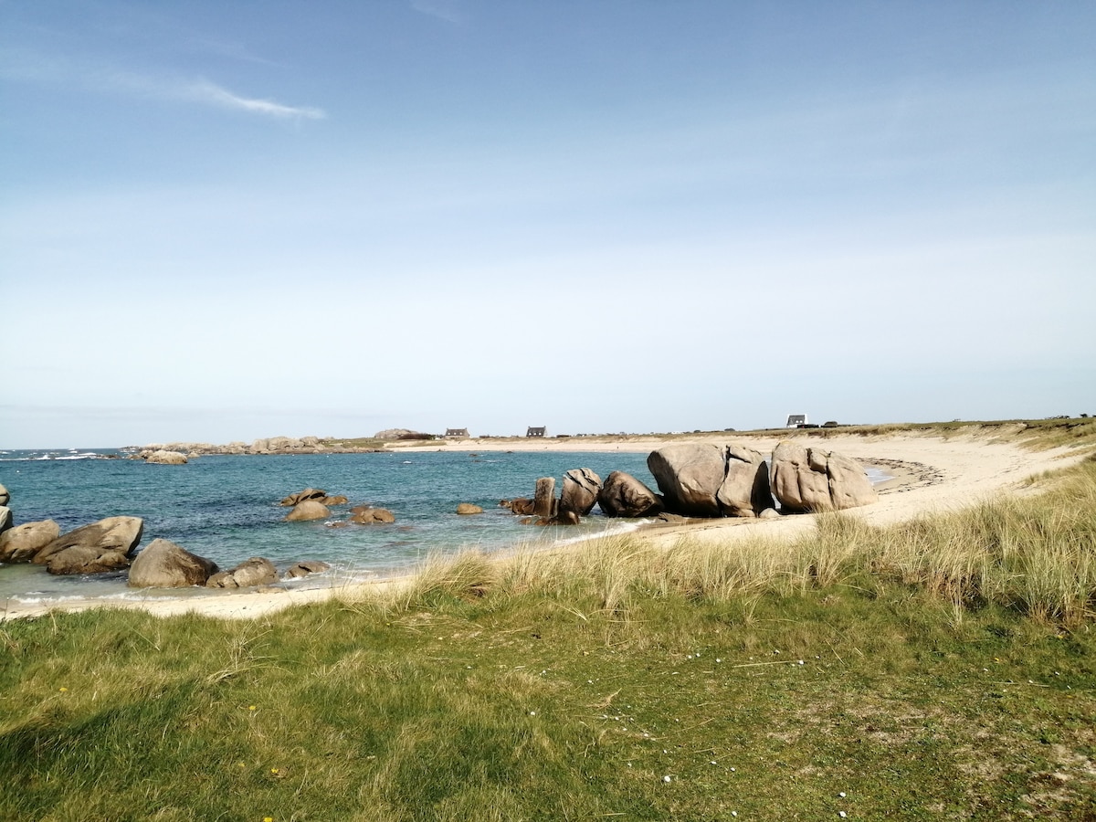 A sandy beach is bordered by smooth rocks and gentle waves. Green grass lines the shoreline, leading toward distant wooden structures on the horizon. The sky is clear with soft blue tones, and the tranquil water reflects the light, creating a serene coastal view.