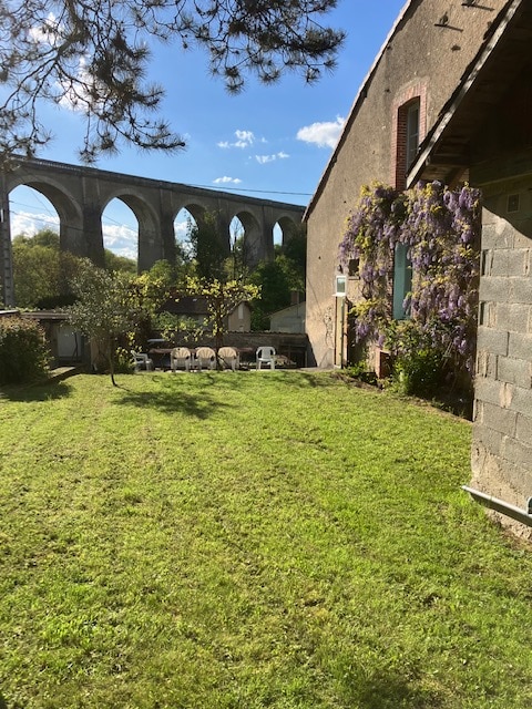 A lush green garden is framed by a historic stone viaduct in the background. Several white chairs are arranged on the lawn, surrounded by trees and greenery. Wisteria vines climb the exterior wall of the building, adding a touch of color to the scene.